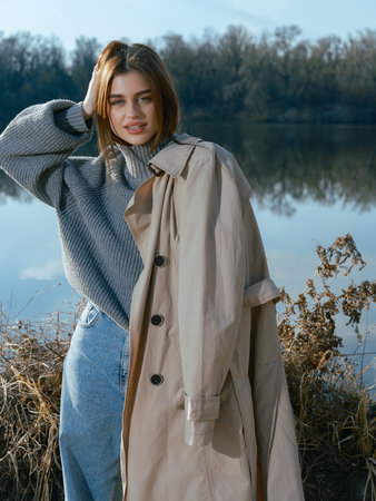 Young woman with trench coat stands by lake with calm waters reflecting the sky, clouds and surrounded by bare trees in early spring. Close-up.の写真素材