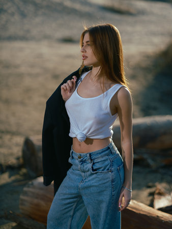 Young brunette woman in white tank top and jeans holding jacket in her hands and standing near logs on sand beach. Close-up.の写真素材