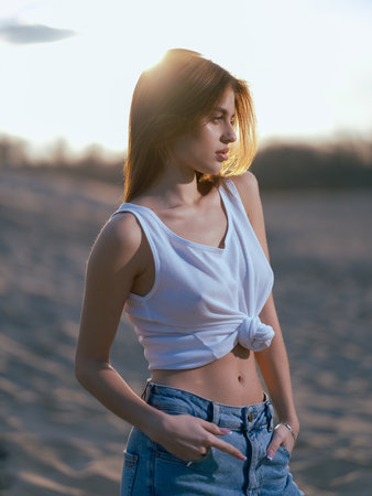 Young brunette woman in white tank top and jeans standing on beach against background of sand and sky at sunset. Close-up.の写真素材