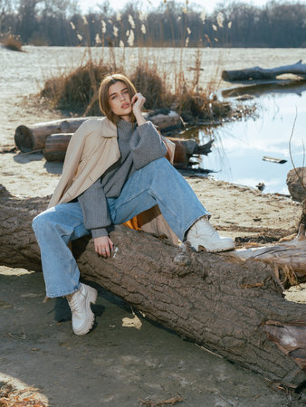 Young woman in trench coat sits on log on beach by lake with calm waters reflecting the sky and clouds in early spring.の写真素材