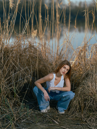 Young brunette woman in white tank top and jeans sitting and relaxing among dry reed against background of lake.の写真素材