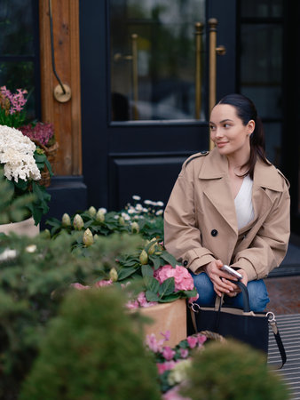 Smiling young woman in jacket and jeans sitting by flower shop, admiring blooming hydrangeas in baskets.の写真素材