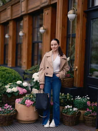 Stylish young woman in jacket and jeans standing by flower shop, surrounded by blooming hydrangeas and cyclamens in baskets.の写真素材