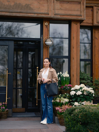 Stylish young woman in jacket and jeans posing by flower shop door, surrounded by blooming hydrangeas, hyacinths and narcissus in baskets.の写真素材