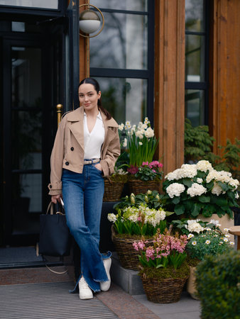 Stylish young woman in jacket and jeans posing by flower shop door, surrounded by blooming hydrangeas, hyacinths and narcissus in baskets.の写真素材