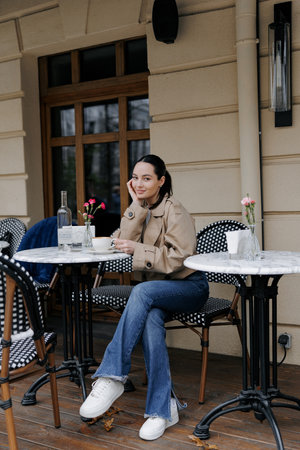 Stylish young woman enjoying coffee at an outdoor cozy cafe. Dressed in jacket and jeans, she sitting near the marble table and smiling.の写真素材