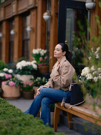 Pensive young woman in jacket and jeans sitting on bench by flower shop, surrounded by blooming hydrangeas in baskets.の写真素材