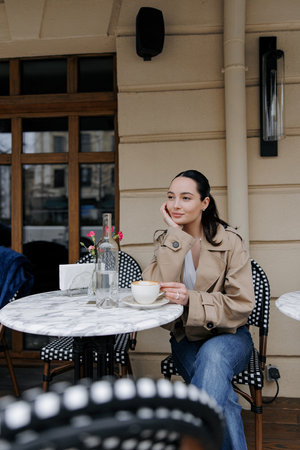 Stylish young woman enjoying coffee at an outdoor cozy cafe. Dressed in jacket and jeans, she sitting near the marble table and relaxing.の写真素材