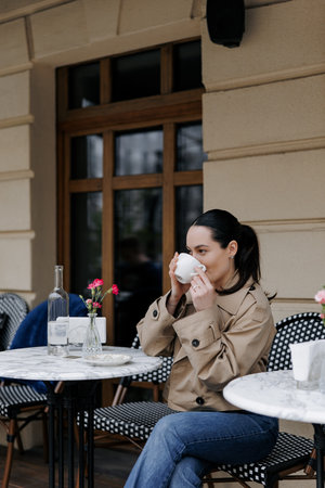 Stylish young woman enjoying coffee at an outdoor cozy cafe. Dressed in jacket and jeans, she sitting near the marble table and drinking.の写真素材