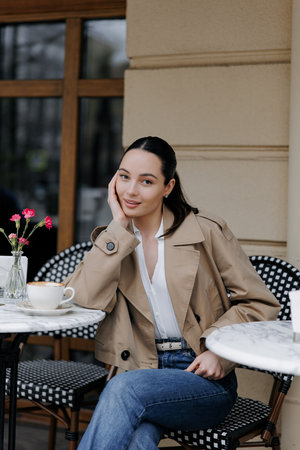 Stylish young woman enjoying coffee at an outdoor cozy cafe. Dressed in jacket and jeans, she sitting near the marble table and smiling. Close-up.の写真素材