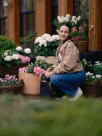 Stylish young woman in jacket and jeans sitting by flower shop, surrounded by blooming hydrangeas and cyclamens in baskets.の写真素材