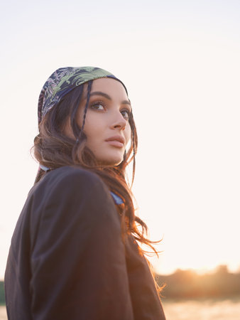 Portrait of young woman in bandana walking among steppe at sunset against sky background. Summer outdoor scene. Backlit. Close-up.の写真素材