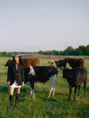 Young woman herding cows on a green pasture in the countryside. Rural summer outdoor scene with livestock, nature and farming life against beautiful landscape.の写真素材