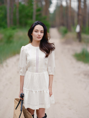 Young woman in white dress holding straw bag, standing on sandy forest path. Calm natural setting with pine trees, peaceful atmosphere, and soft daylight. Close-up.の写真素材