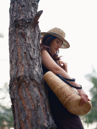 Young woman in brown dress and straw hat holding straw bag, standing against background of pine trees and sky. Bottom view. Calm natural setting, peaceful atmosphere, and soft daylight. Close-up.の写真素材