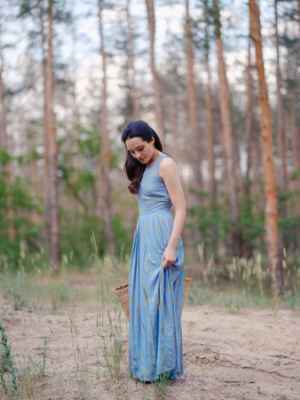 Young woman in blue dress holding straw bag, standing on sandy forest path. Bottom view. Calm natural setting with pine trees, peaceful atmosphere, and soft daylight.の写真素材