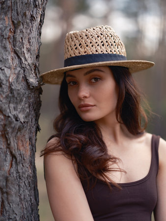 Portrait of young woman in brown dress and straw hat standing against blur background of pine tree forest. Close-up. Calm natural setting with pine trees, peaceful atmosphere, and soft daylight.の写真素材