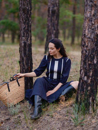 Young woman in dark purple dress and boots sitting among pine tree trunks in forest next her straw bag. Calm natural setting with pine trees, peaceful atmosphere, and soft daylight.の写真素材
