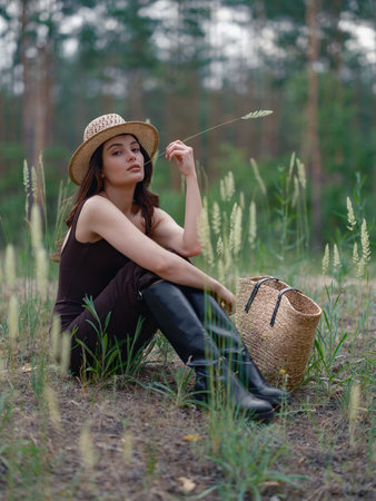 Young woman in brown dress and straw hat sitting and resting in forest next her straw bag. Calm natural setting with pine trees, peaceful atmosphere, and soft daylight.の写真素材