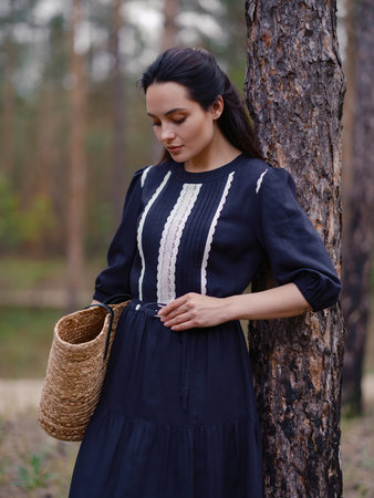 Young woman in dark purple dress holding straw bag, standing among pine tree trunks in forest. Close-up. Calm natural setting with pine trees, peaceful atmosphere, and soft daylight.の写真素材
