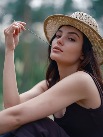 Portrait of young woman in brown dress and straw hat sitting and relaxing against blur background of forest. Close-up. Calm natural setting, peaceful atmosphere, and soft daylight.の写真素材