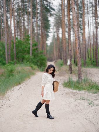 Young woman in white dress and black boots holding straw bag, standing on sandy forest path. Calm natural setting with pine trees, peaceful atmosphere, and soft daylight.の写真素材