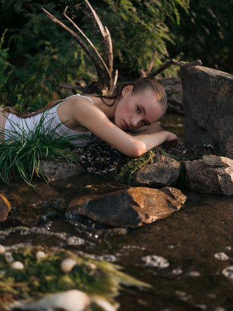 Young wet woman lies by shallow forest stream among rocks and grass. Peaceful natural setting, soft light, and calm mood. Summer day, nature connection, and outdoor relaxation.の写真素材