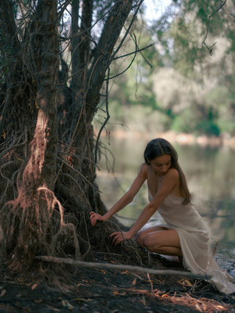 Young woman in white dress sits and rests by forest lake among old tree roots. Tranquil atmosphere in nature. Fairytale, magical, mysterious and mystical forest. Close-up.の写真素材