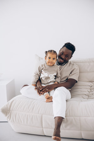 Young african man sitting on sofa and cheerfully playing with his biracial toddler daughter. Concept of interracial family.の写真素材
