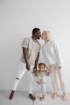 Loving biracial family holding hands their daughter sharing a tender moment of kiss against a white...