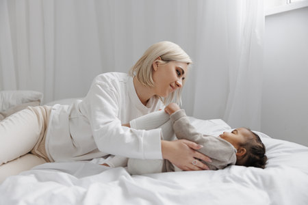 Happy mother and her biracial baby daughter playing together on bed. Concept of interracial family.の写真素材