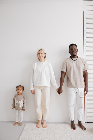 Happy, smiling biracial family of three stands against a white wall in a simple setting. Concept of...