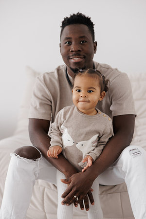 Happy father is holding his sweet biracial daughter on his lap, both looking at the camera. Concept of interracial family.の写真素材