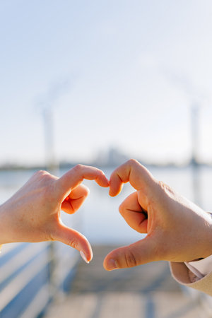 Close-up of loving couple hands forming a heart shape with fingers on waterfront pier with blurred cityscape view. Romantic gesture symbolizing love and connection.の写真素材
