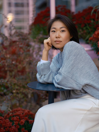 Close up portrait of young Asian woman in light sweater looking at camera. She is sitting at a table...