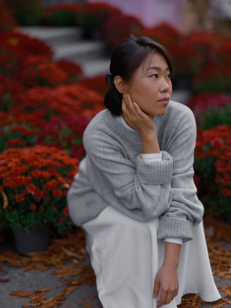 Young Asian thoughtful woman crouching or squatting on the pavement and looking away among vibrant red chrysanthemum flowers and fallen leaves against urban autumn setting.の写真素材