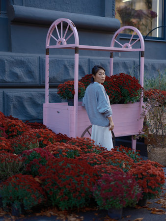 Stylish young Asian woman standing and turning back next to a pink wooden flower cart. Vibrant red chrysanthemum flowers create a beautiful urban autumn scene.の写真素材