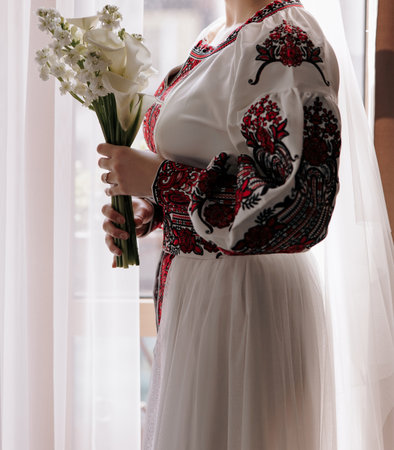 Ukrainian bride in traditional red and white embroidered Vyshyvanka dress, holding elegant calla lily bouquet near window with curtains. Authentic ethnic fashion and cultural traditions in Ukraine.の写真素材