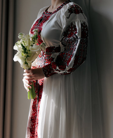 Side view of Ukrainian bride in red and white embroidered Vyshyvanka dress, holding elegant calla...