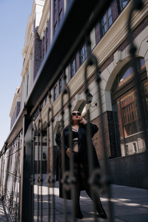 Full-length low-angle shot of stylish plus size woman in black lingerie and leather trench coat. Confident curvy model poses near building and railing, promoting body positivity.の写真素材