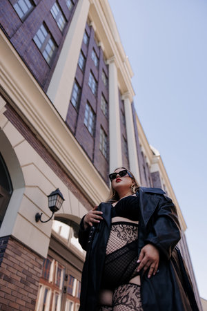 Extreme low-angle view of stylish plus size woman in black lingerie and leather trench coat. Confident curvy model poses near imposing urban architecture, promoting body positivity.の写真素材