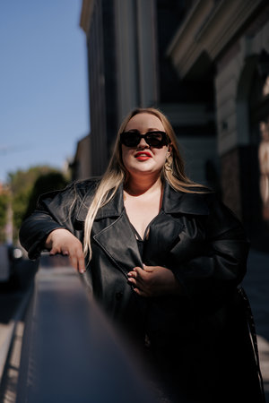 Close-up portrait of stylish plus size woman in black leather trench coat and sunglasses. Confident curvy model leaning on railing, promoting body positivity and urban fashion.の写真素材