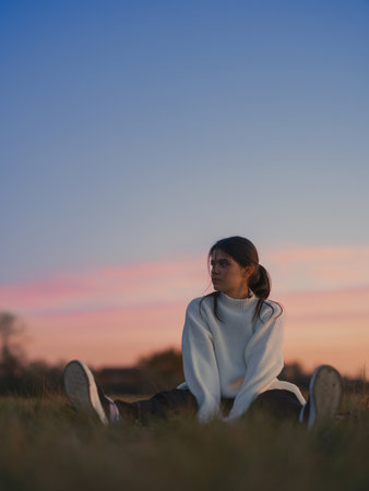 Low angle shot of young woman sitting on ground in field with vivid pink and blue sunset sky. She...