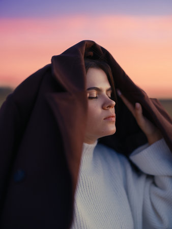 Emotional portrait young woman with closed eyes covering head with coat against vibrant pink and purple sunset sky feeling peaceful moment.の写真素材
