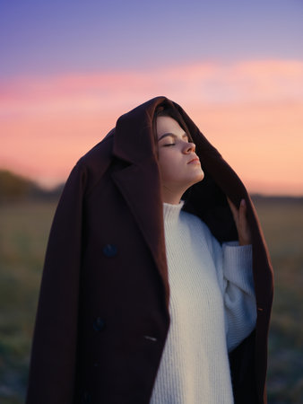 Emotional portrait young woman with closed eyes covering head with coat against vibrant pink and purple sunset sky feeling peaceful moment.の写真素材