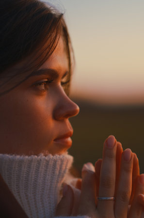 Extreme close-up profile portrait of young woman with hands folded during warm sunset light. She wears white sweater and expresses thoughtful, hopeful, and serene mood outdoors.の写真素材