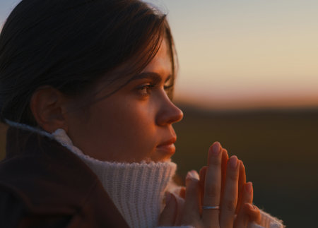 Close-up profile portrait of young woman with hands folded during warm sunset light. She wears a white sweater and expresses thoughtful, hopeful, and serene mood outdoors.の写真素材