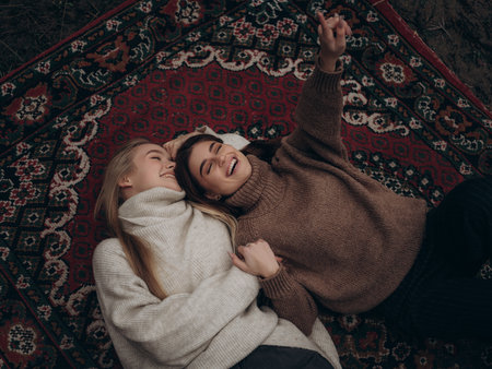 Happy young lesbian couple laughing frankly lying on ornate carpet looking up pointing finger. Fun, relaxation, and shared happy moment in diverse LGBT relationship.の写真素材
