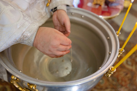 Close-up of priest's hands holding small silver cross necklace over water in large baptismal font...