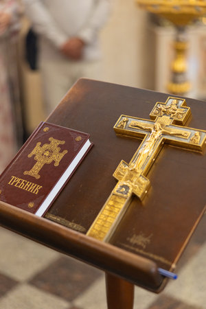 Close-up of golden Crucifix and burgundy Trebnik (service book) on wooden lectern in Orthodox church interior. Symbolizes religious service, faith, prayer, and Christian tradition.の写真素材